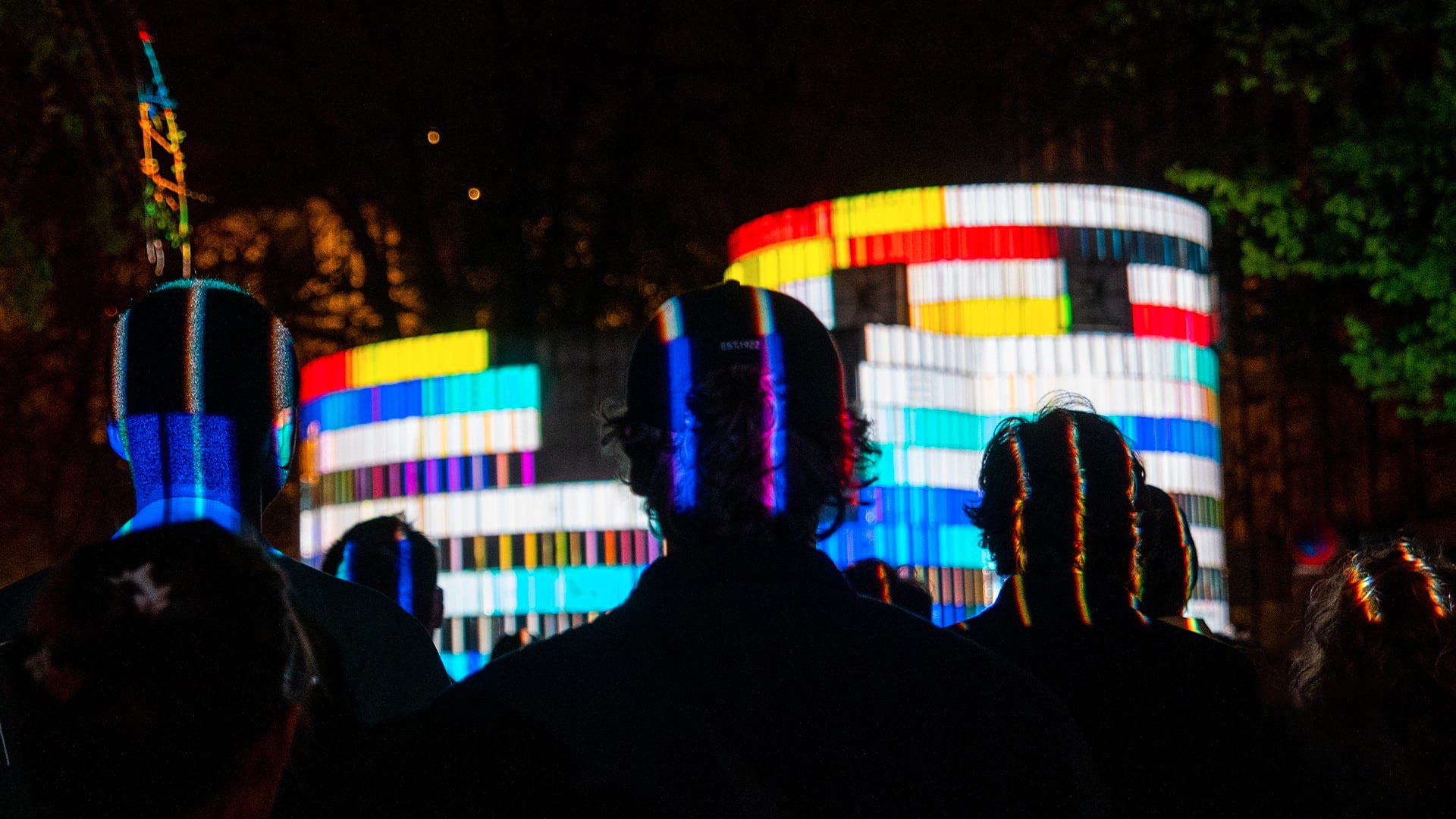 874 projection mapping on the curved concrete parking ventilation shaft at Nouveau Siècle in Lille at night, hard blocks of red, yellow, cyan and white light banded around the structure with an audience silhouetted in front